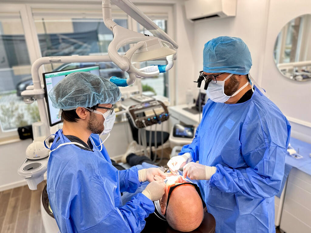„Лекар и асистент извършват орална хирургия с увеличителни очила и дигитално оборудване.“